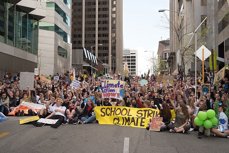 A crowd of young protesters sit in a city street with signs stating “Climate Action Now” and “School Strike.”