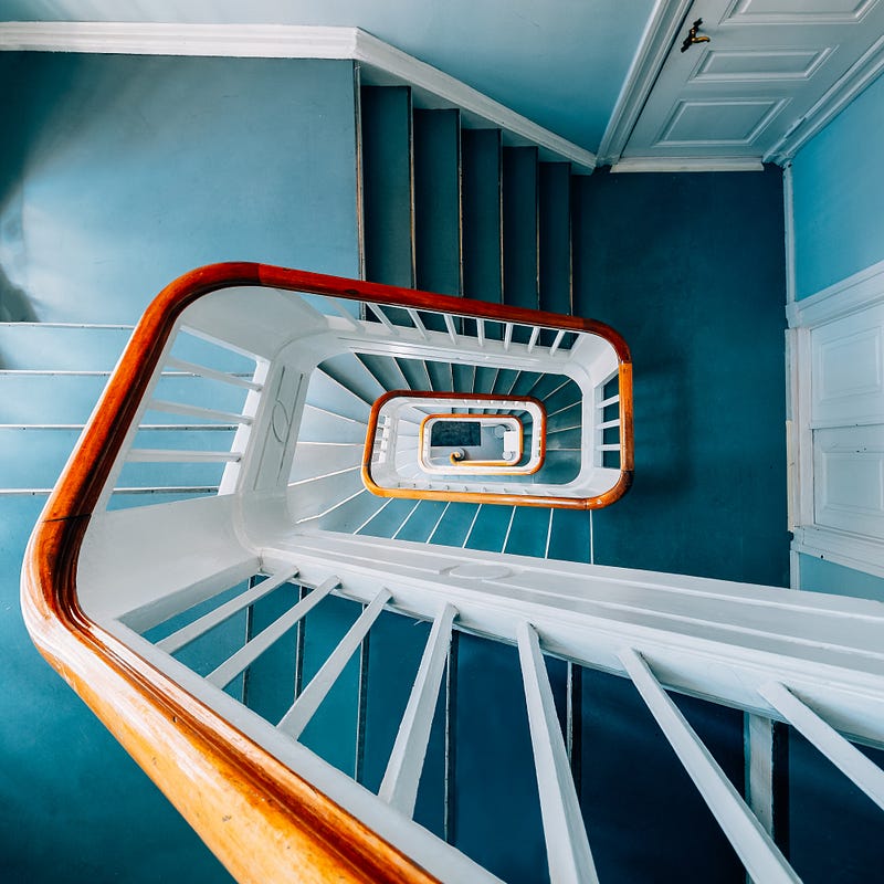 This elegant spiral staircase showcases a timeless architectural design with a smooth wooden handrail and crisp white balusters set against soothing blue walls. The geometric symmetry and vertical perspective create a striking visual rhythm, highlighting craftsmanship and structural beauty. Ideal for classic and heritage residential interiors, this staircase design blends functionality with aesthetic appeal, making it a focal point that enhances spatial flow and architectural character.