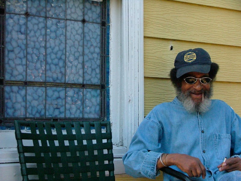 Photo By Rashida Simmons of an older Black man with grey bears, with his arms on the chair, a smiling face, and a stick in his mouth. He is wearing a navy blue baseball cap, wire framed glasses, and a light blue denim shirt sitting by a window.