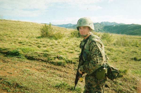 Young U.S. Marine Cpl. Chad Eric Oligschlaeger in uniform holding a rifle during deployment, standing in a grassy, mountainous landscape