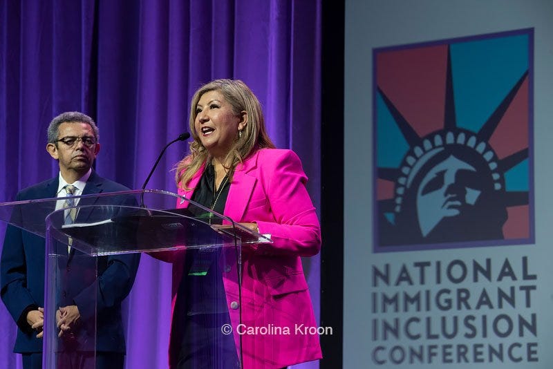 NPNA Executive Director Nicole Melaku and NPNA Board Co-Chair Gustavo Torres speaking in front of a podium on a stage at the 2022 National Immigrant Inclusion Conference (NIIC) with the NIIC logo in the background. Photo Credit: Carolina Croon