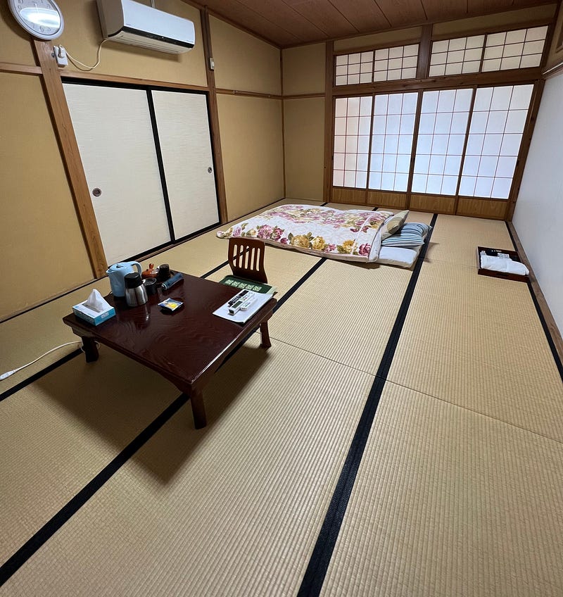 Large tatami mat room with futon bed laid out and low table with floor chair.