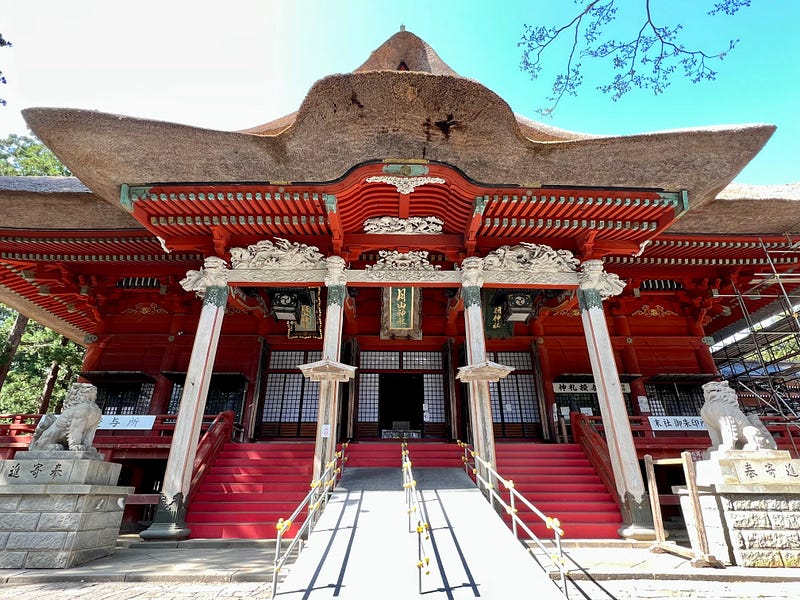 Red shrine with thick thatched roof. The shrine is guarded by 2 komainu lion dog statues and is decorated with carvings.