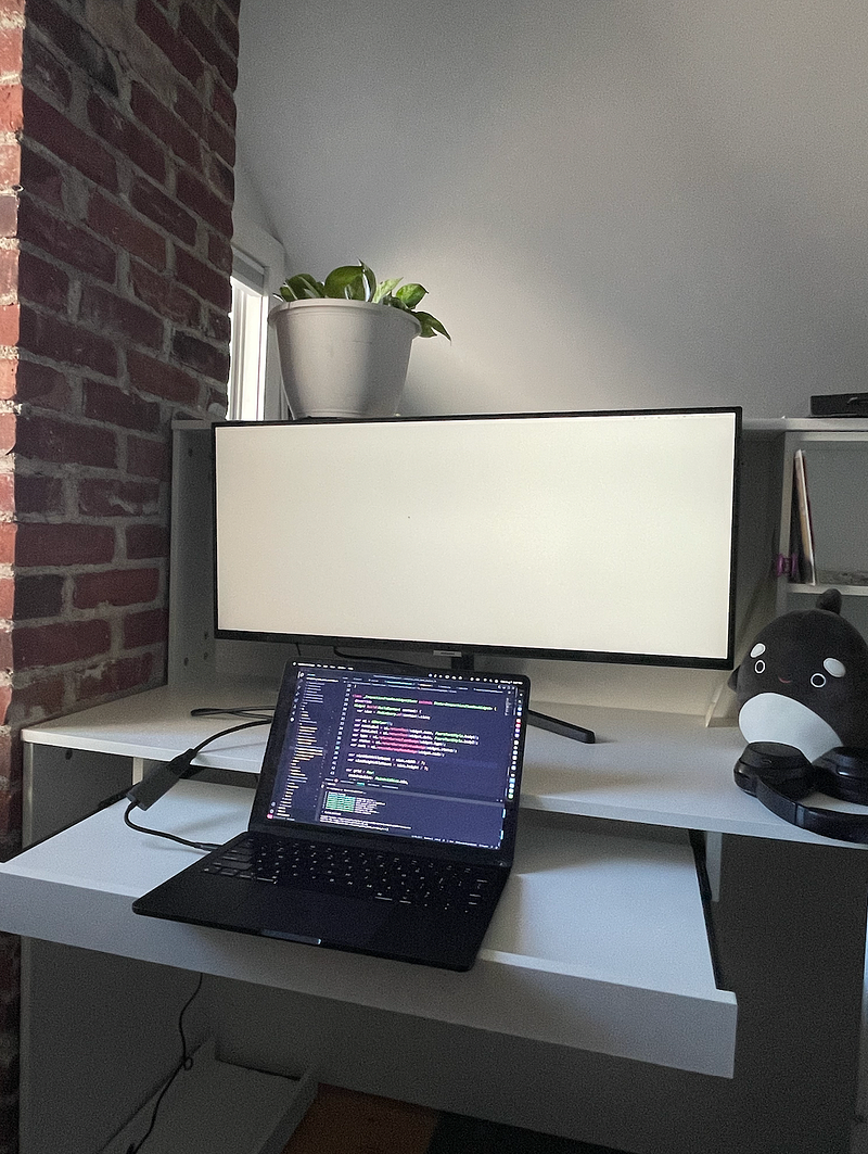 My desk with a large monitor, a laptop with website source code, and my Orca squishmallow.