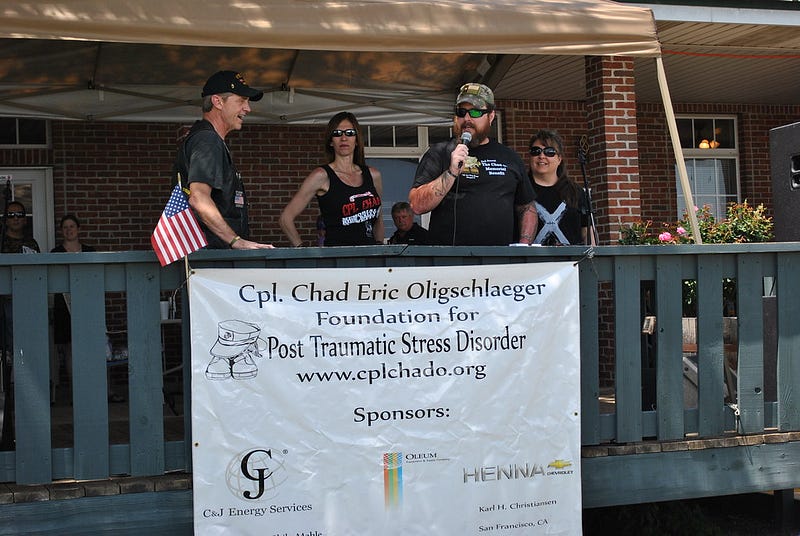 People speaking at a Chad O Foundation PTSD awareness fundraiser, standing behind a banner honoring Cpl. Chad Eric Oligschlaeger