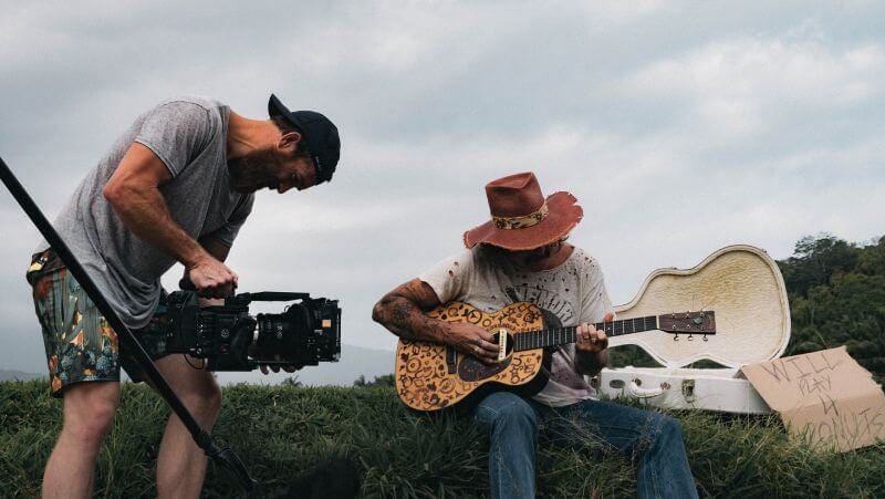 A man shooting a video of a woman sitting while holding an acoustic guitar.