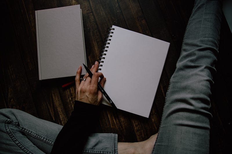 A woman sitting on the floor with a blank page in an open notebook, ready to journal with a pen in hand.