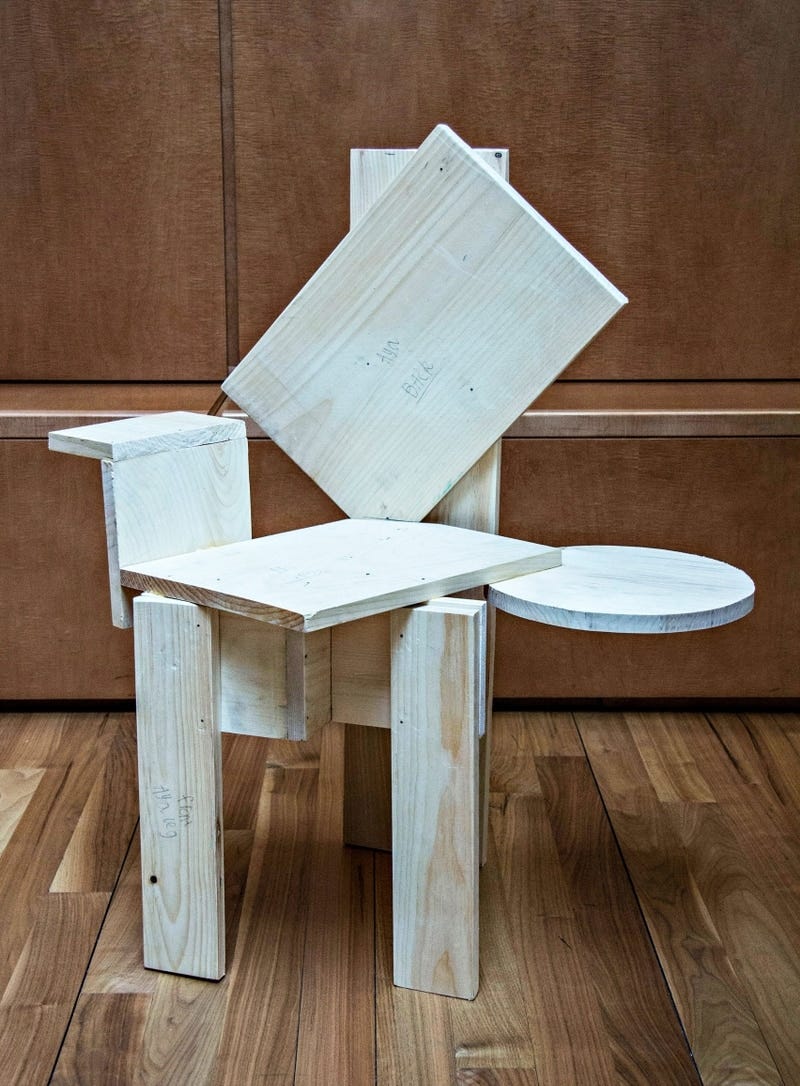 A chair made of wooden panels. The seat has been turned forty-five degrees from being in line with the legs, with an arm rest on its right side in line with the seat. On the left, instead of an arm rest, there is a small circle extending at the height of the seat. The rectangular back of the chair is also rotated to the left by forty-five degrees.