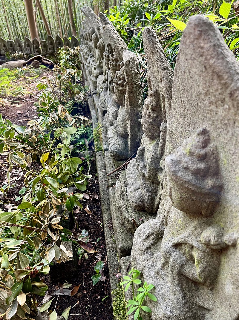 Buddhist statues lined up amid weeds and in front of a bamboo grove.