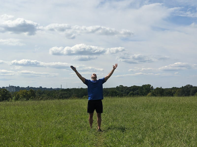 A Native American man wearing t-shirt and shorts, standing in an open field with arms open towards peaceful blue skies and fluffy white clouds