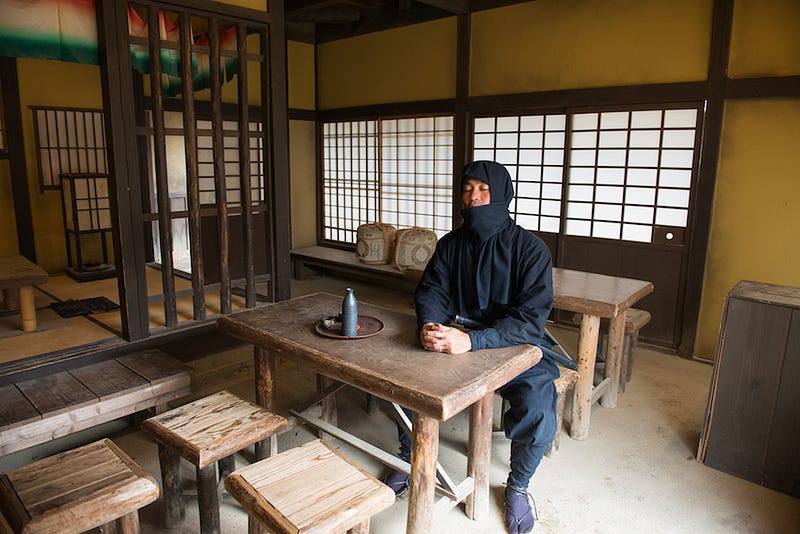 A lone ninja sits at a table drinking sake
