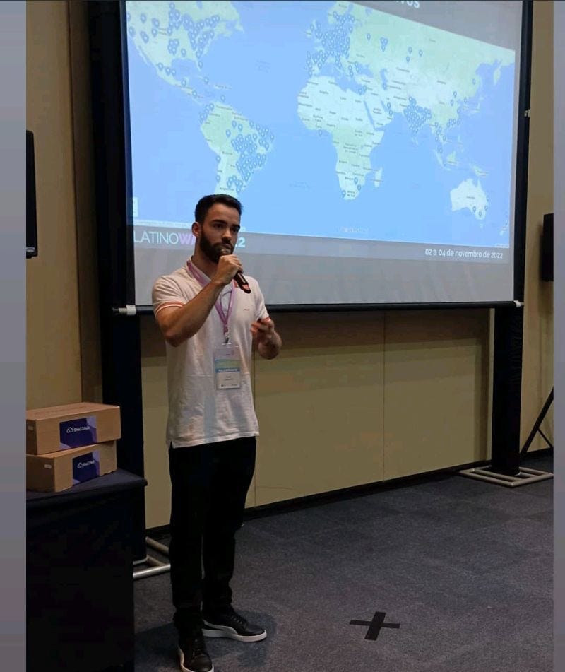 palestrante Gesse Silva de camisa polo branca e calça jeans preta, em frente a uma tele onde está reproduzido o mapa mundi com os mais de 200 mil devices que utilizam shellhub espalhados ao redor do mundo.