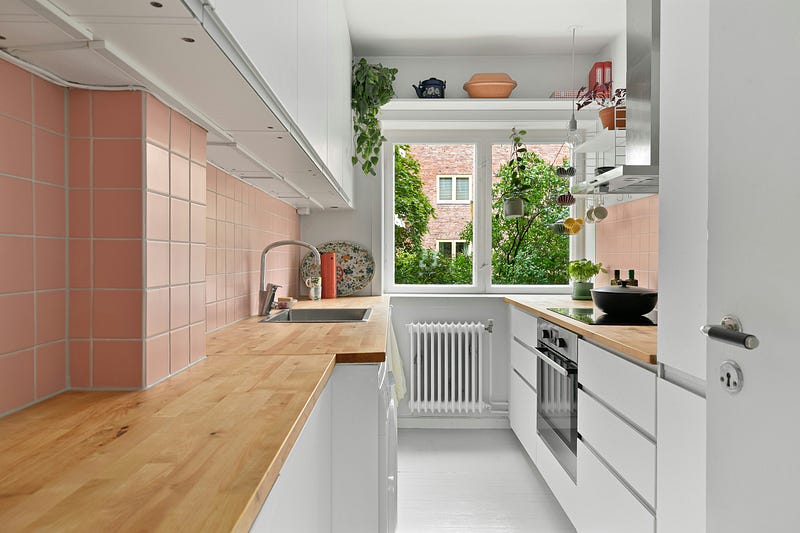This modern parallel kitchen design showcases a clean and efficient galley layout with white cabinetry and warm wooden countertops. Soft pastel tile backsplash adds subtle color and character, while natural light from the large window enhances brightness and ventilation. Integrated appliances, smart storage solutions, and indoor plants create a fresh, functional, and inviting cooking space — ideal for compact homes and contemporary apartments.