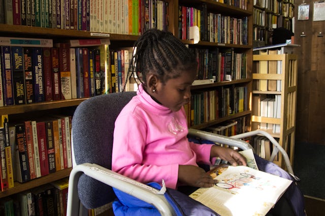 Picture of a young Black girl sitting in a chair in front of a large bookshelf reading a book.