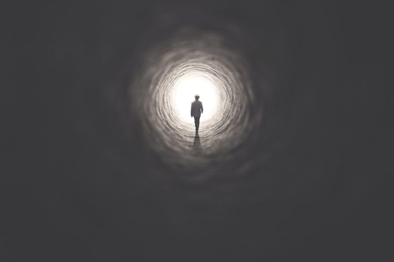 A man walks down a tunnel towards a point of light in the distance.