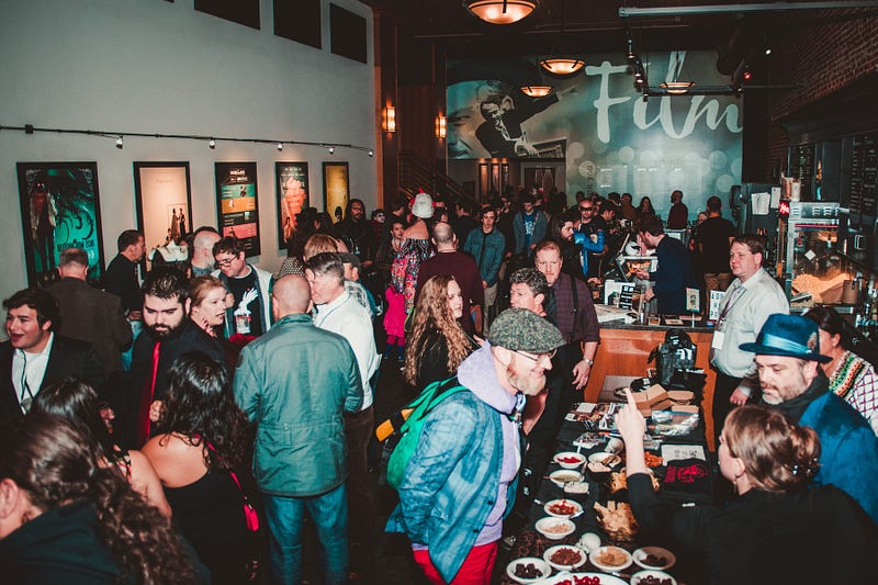 Over fifty people fill a reception hall with horror posters on the walls and a food table.