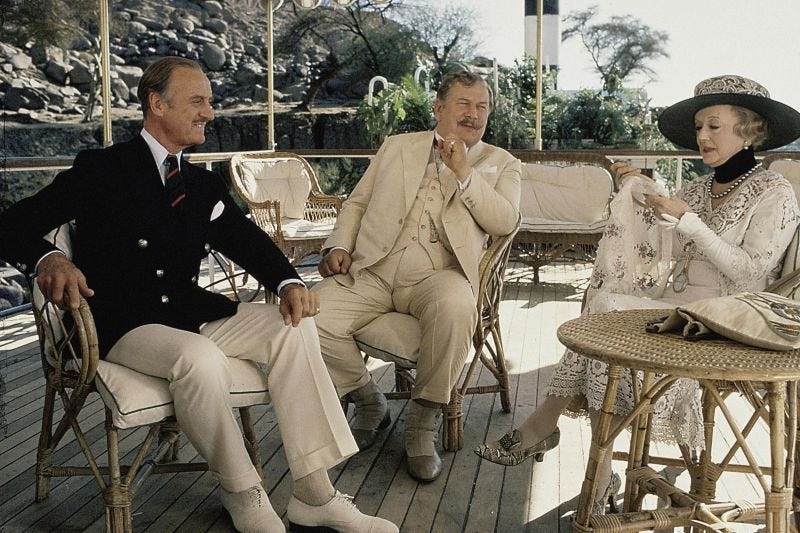 David Niven (as Colonel Race), Peter Ustinov (as Hercule Poirot) sit with Bette Davis (as Marie Van Shuyler), who is doing embroidery. They are on a boat in the 1978 movie adaptation of Death on the Nile