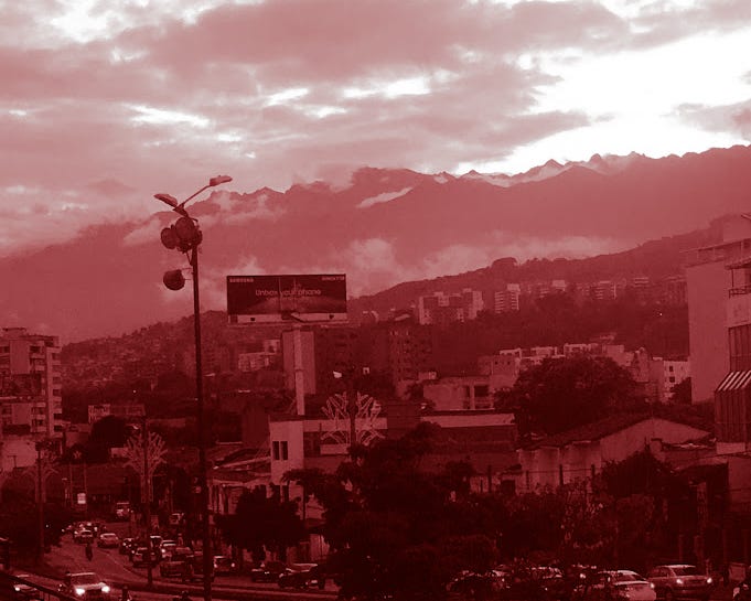 Colorized red photo of city in in the foreground and mountains behind with an ominous cloudy sky with bright sun shining through.