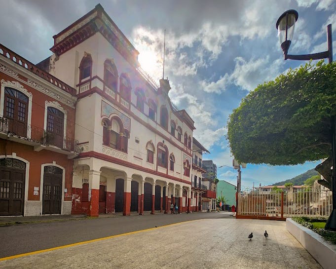 Looking down a street lined with colonial style red and whit buildings in the background are green mountains. Abover a sunny blue sky with puffy scattered clouds.