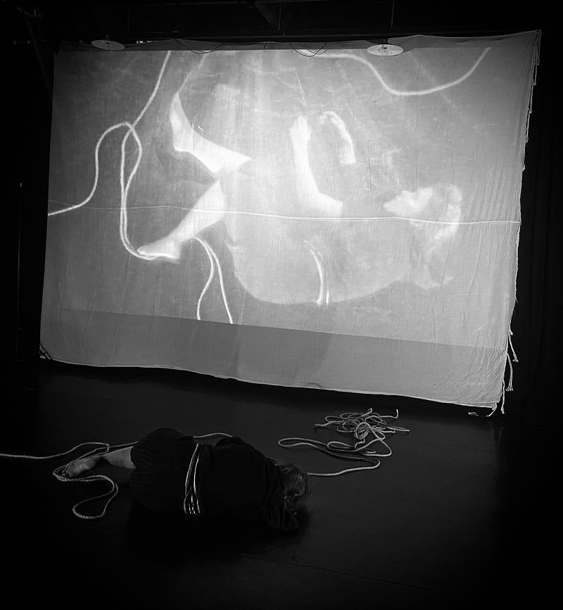 A dancer lies on the floor with a piece of rope coiled around her. An image of her is captured by an overhead camera and this is projected onto a screen behind her. In the image it looks like she and the rope arefalling through deep water.