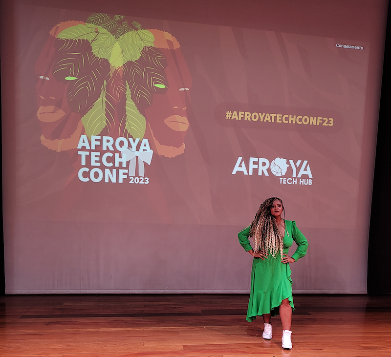 A imagem apresenta a Andreza Rocha, idealizadora do evento. Ela é uma mulher negra, com uma roupa verde, cabelo longo e, ao seu fundo, há um painel da conferência Afroya Tech Conf.