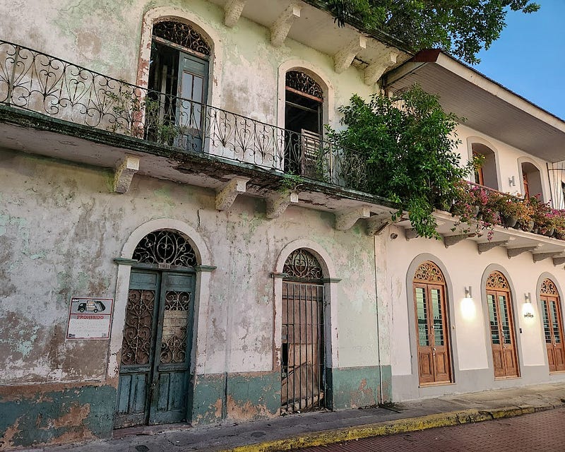 Two attached building fronts, nearly identical except that the one on the right is well preserved, clean, painted, adorned with plants and decorative light fixtures, while the one on the left has faded and chipped paint, broken doors and glass, boarded windows and no decoration.