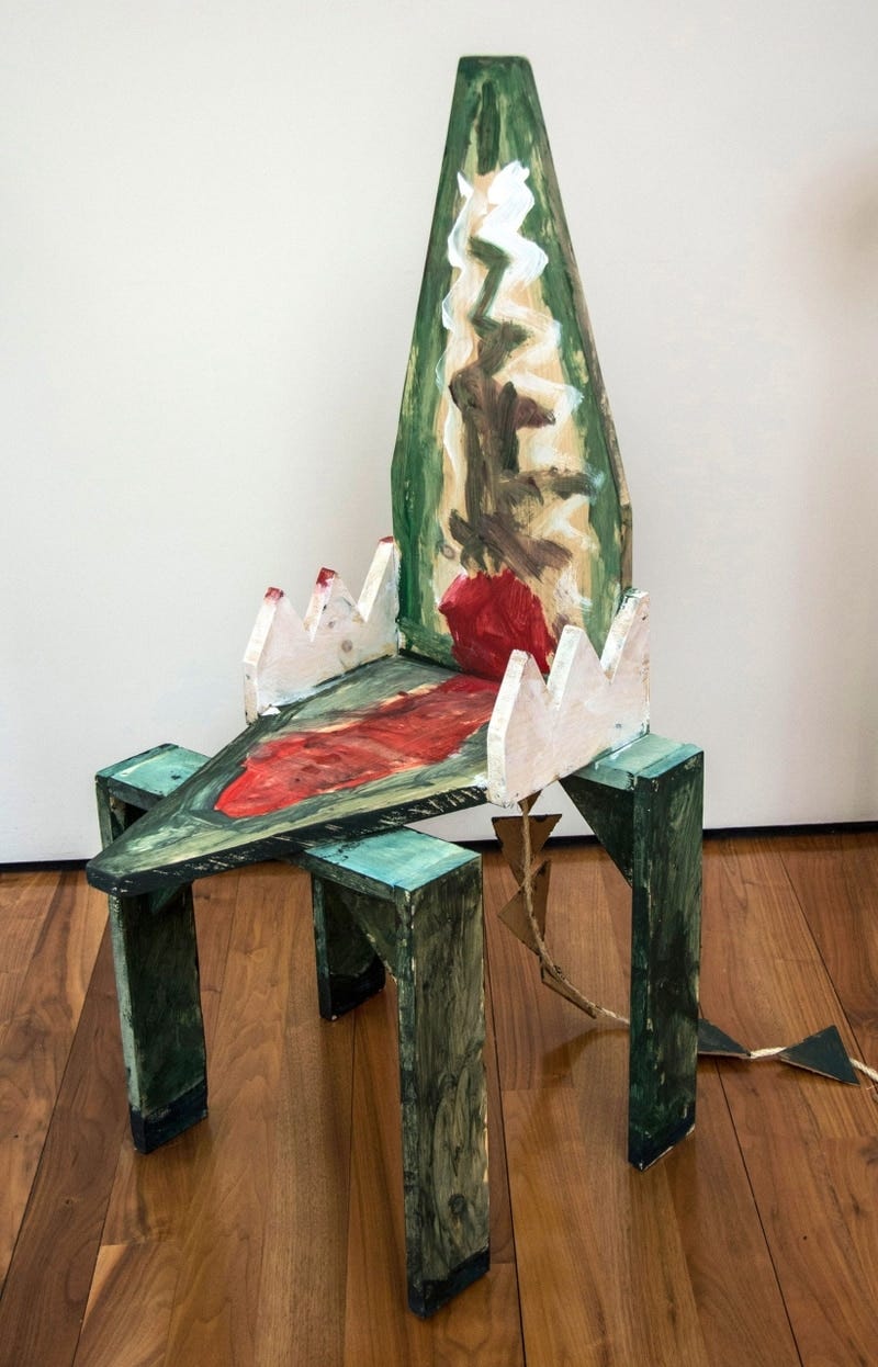 A chair designed to look like a crocodile’s mouth, painted mostly green. There are four rectangular chair legs, and the mouth is open to form the seat and back, with a painted red tongue. The arm rests are each made of three jagged white teeth. The chair also has a tail made of a string with triangle pieces along it, stuck to the back end of the chair.