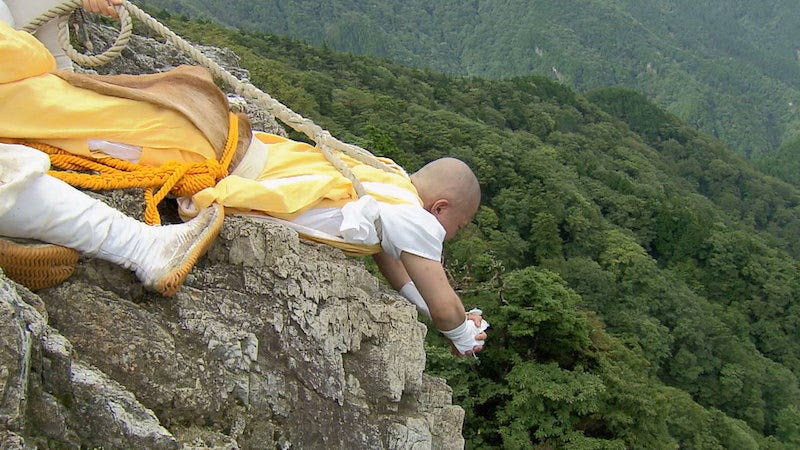 A yamabushi holds an mountain asceticism trainee over a cliff by nothing by a rope on Nara Prefecture’s Mt. Omine