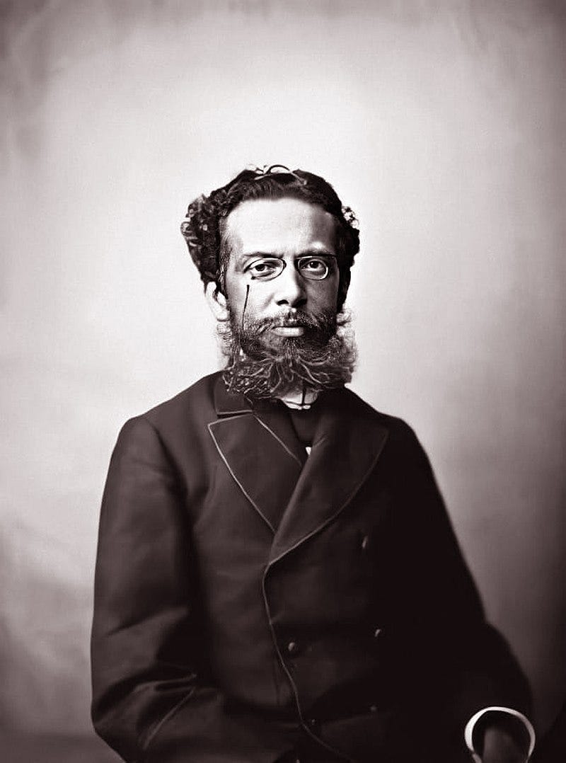 archival photo of Machado de Assis, a mixed-race man with a long, bushy beard and short hair. He wears very small eyeglasses and a double-breasted coat. He has a serious look on his face, as people in old photos often do.
