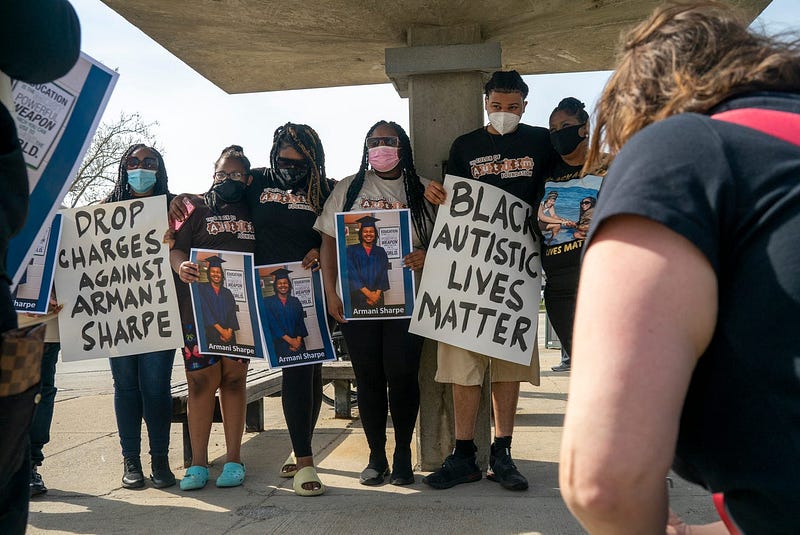 Photo from protest showing 5 African American individuals holding signs and photos. Signs read “Drop Charges Against Armani Sharpe” and “Black Autistic Lives Matter”