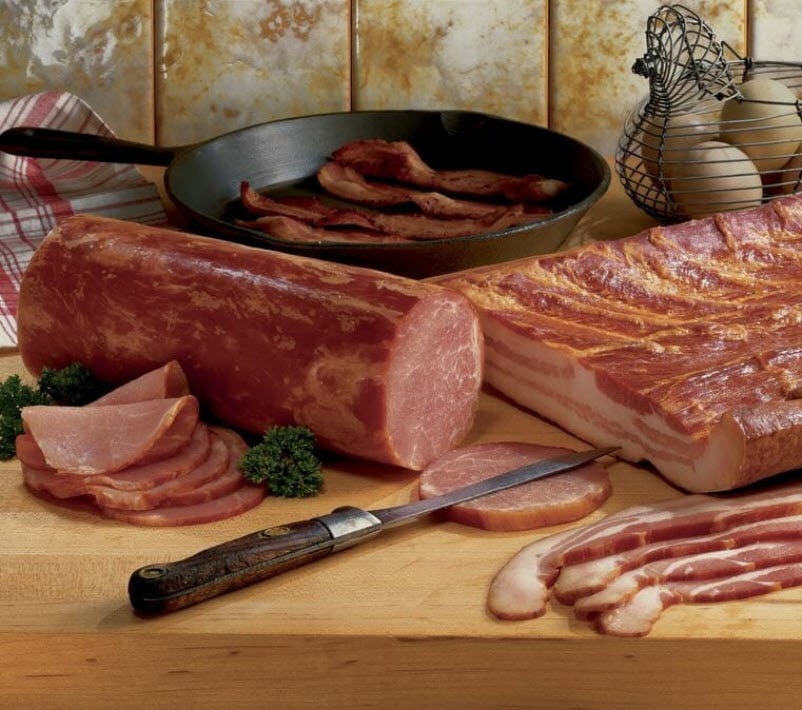 A Canadian bacon log and slices next to a rack and strips of bacon, with a cast iron pan and a wire basket of eggs.