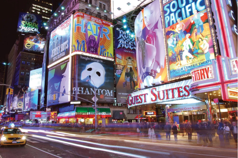 A bright city-scape street, many buildings are having posters for musicals such as “Wicked,” “Phantom of the Opera,” “Hair,” and others that are not readable. There are crowds of people walking down the streets, some entering the building with the sign “Guest Suites.” There is a taxi in the corner waiting and the entire picture is in bright and glowing colors.
