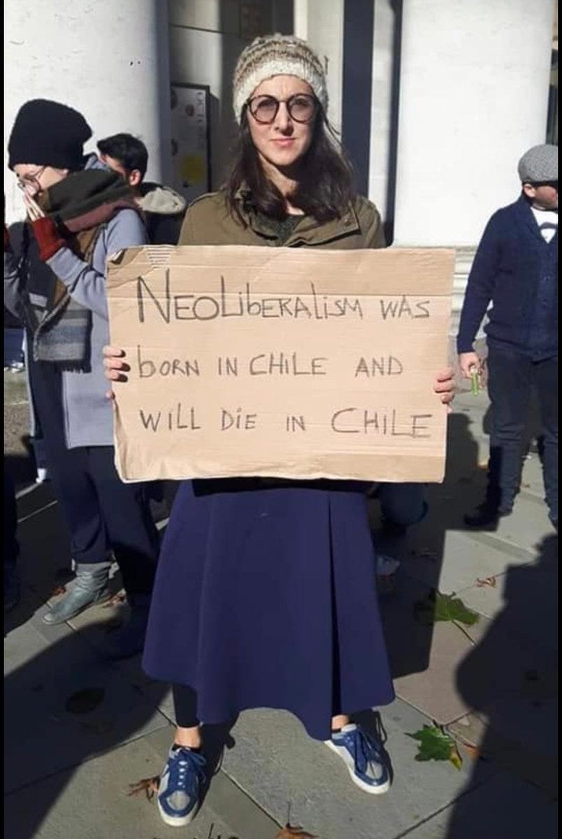 photo of woman holding cardboard sign saying “neoliberalism was born in Chile and will die in Chile”