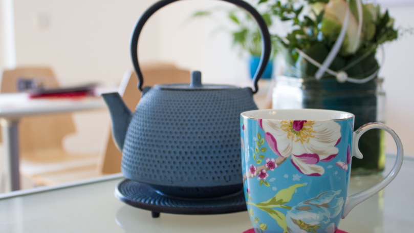 A blue tea kettle sits on top of a darker blue hot plate on a counter. In front of it is a blue mug painted with colorful floral designs.