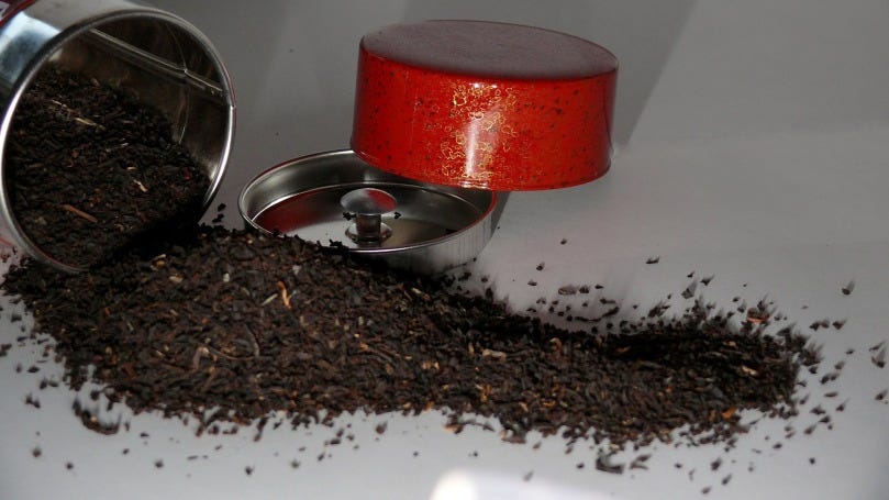 Loose black tea leaves spilling out of a metal container and onto a white table top. The red cap of the container and the metal lid to create an airtight seal with the container rest against each other in the background.