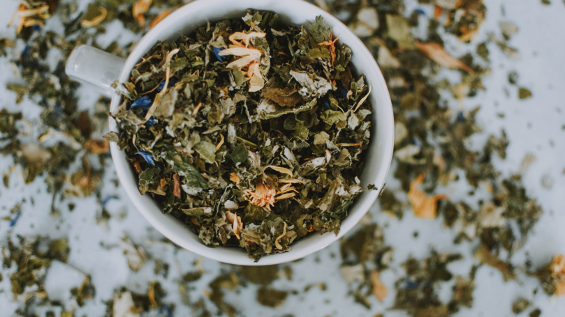 A top down shot of loose green tea leaves and a variety of colorful flower petals in a white mug. The remaining leaves are scattered on the table around the mug.