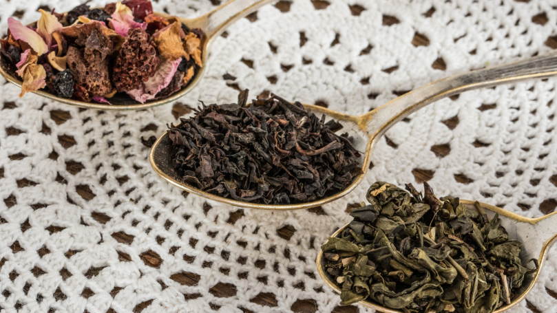 An assortment of loose tea leaves in three golden spoons in front of a white doily. The topmost spoon contains a scoop of herbal tea that includes colorful flower petals. The middle spoon contains black tea leaves. The bottom spoon contains green tea leaves.