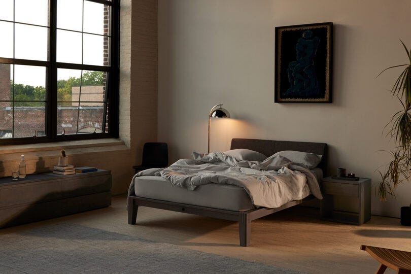 Modern bedroom with a large window, a wooden platform bed with white bedding, potted plants, and abstract art on the wall at dusk.