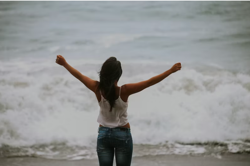 Woman standing with arms open in front of the ocean with waves crashing toward her