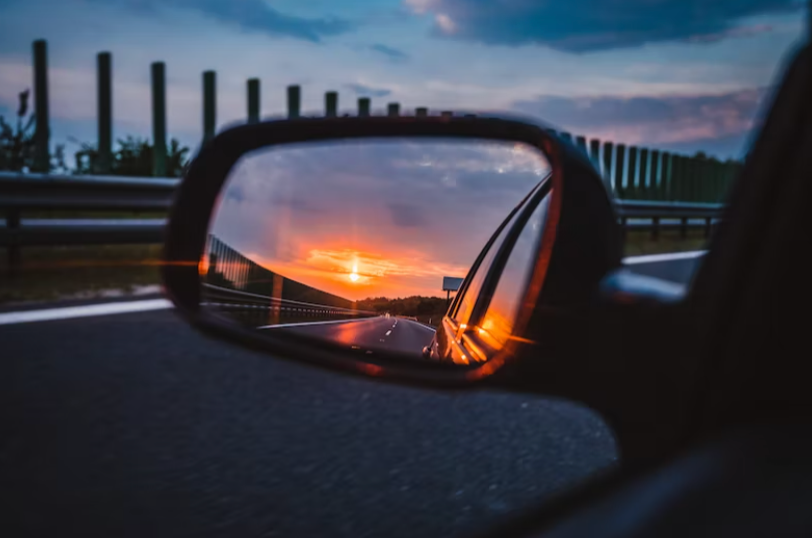 Imagem contendo ao fundo uma estrada com céu azul. ao centro, o foco de um retrovisor de carro reflete o por do sol
