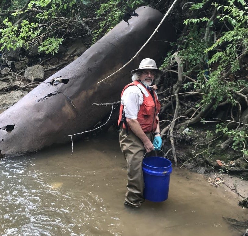 A man in a Fish and Wildlife Service uniform, orange life jacket, and sun hat stands in a shallow river holding a blue bucket, with a large rusted pipe behind him.