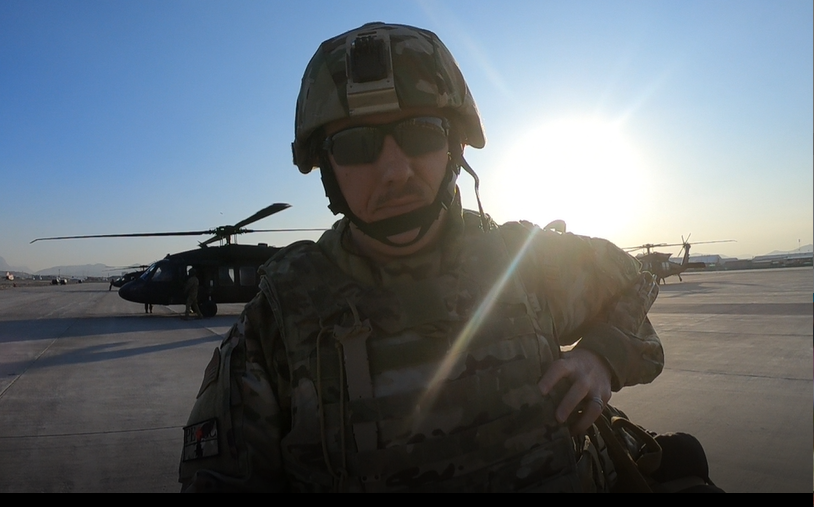 Christopher Sage in military uniform with military helicopters and the sun behind him.