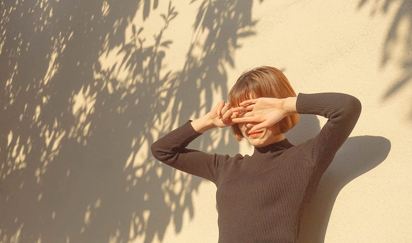 Woman with bob and bangs holds hands face outward over her eyes. She stands against a cream background decorated with gray leaf shadows.