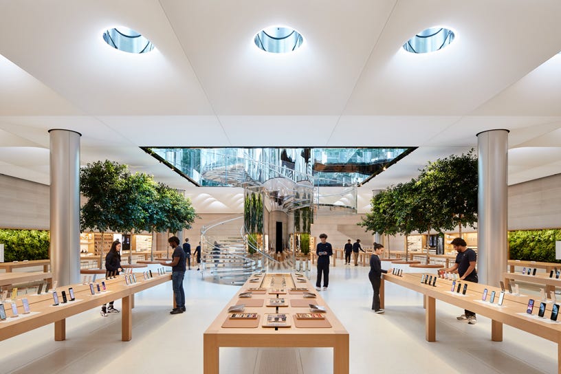 Modern tech store interior with display tables, staff, and a central glass staircase.