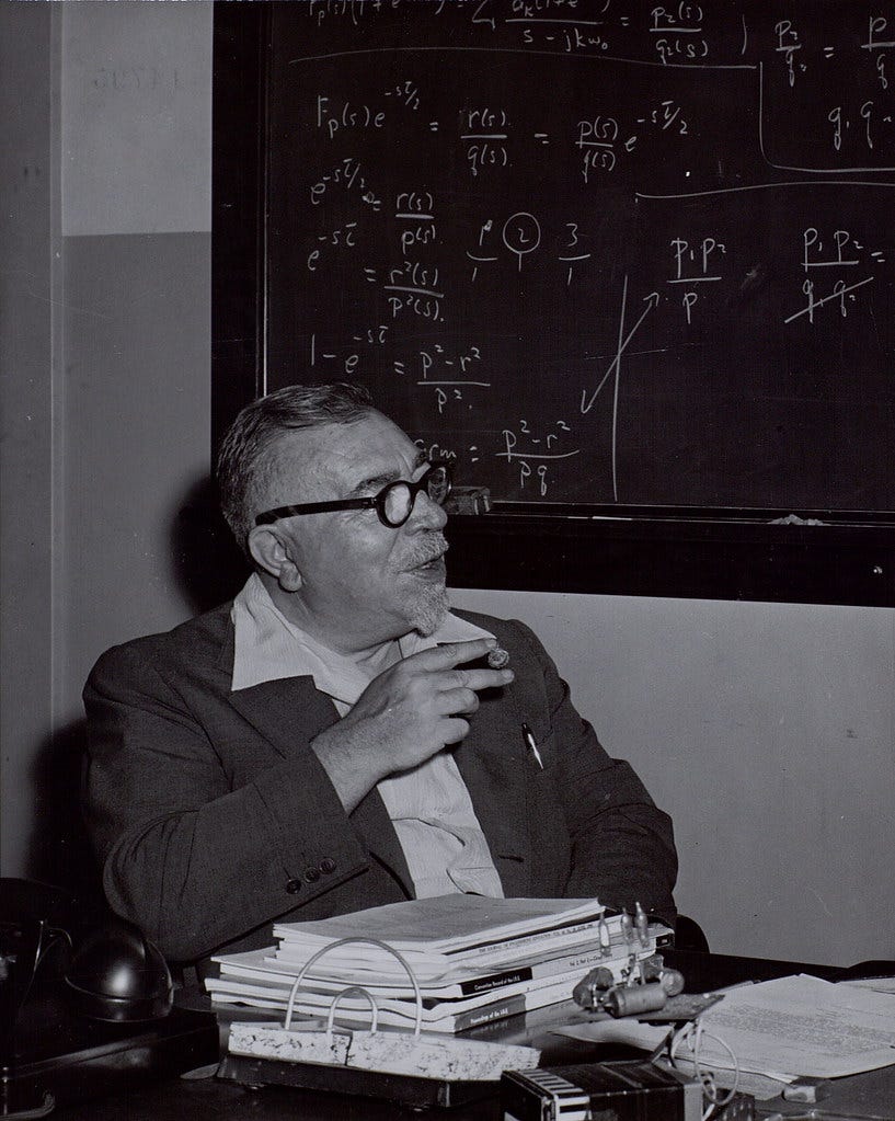 A black-and-white photo of Norbert Wiener, sat in front of a chalkboard, with complicated equations written all over it.