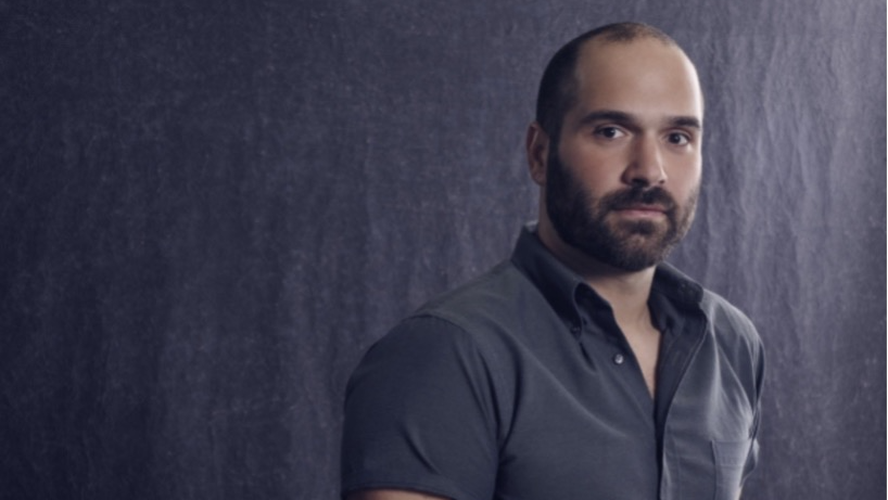 A man with short dark hair and a close-cropped beard wearing a gray shirt looks at the camera