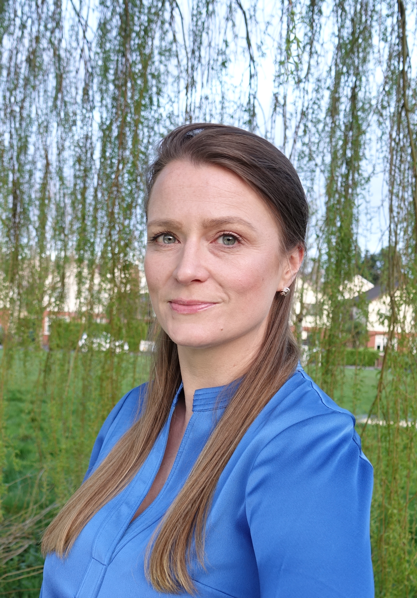 A woman with dark blonde hair wearing a blue top. She is standing in front of a weeping willow on a green.