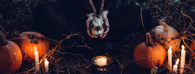 Imagem que faz referência a um ritual de bruxaria, com duas mãos segurando o crânio de um animal, velas e abóboras ao redor