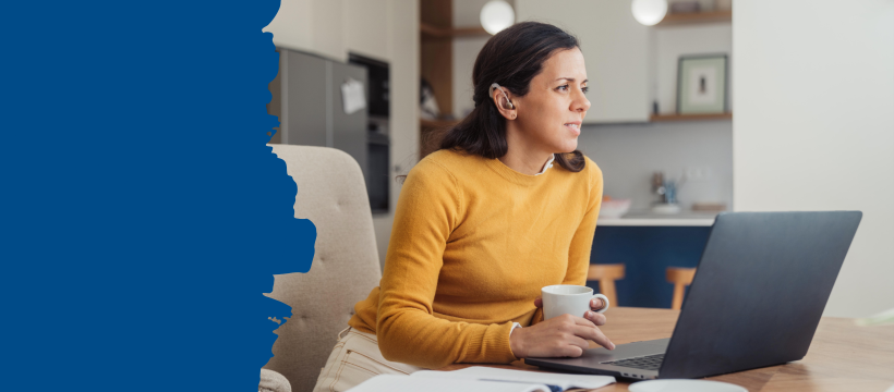 Woman wearing a yellow jumper, holding a mug and sitting at a laptop. A hearing aid is visible in her right ear.