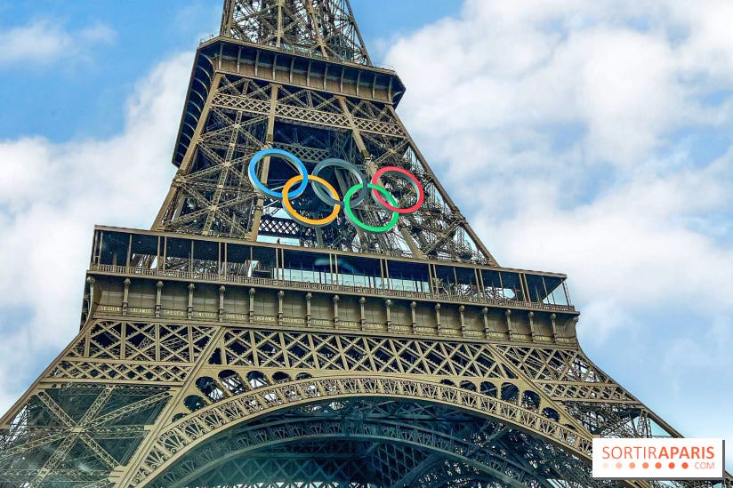 Foto tirada no ângulo de baixo para cima da Torre Eiffel. Ao centro, é possível ver o símbolo das olimpíadas, que é formado por cinco argolas que se entrelaçam em duas fileiras. Na primeira, três argolas: uma azul, uma preta e uma vermelha. Na segunda, duas: uma amarela e uma verde.